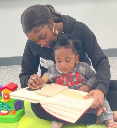 A little boy with dark skin and hair sits on his mothers lap playing with the tail of a wolf from a tactile picture book, Little Red Riding Hood. Both are concentrating hard on the page.