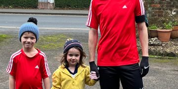 A white man in his 30's, wearing running gear stands on a driveway holding the hand of his toddler daughter, also in running gear. Holding the girl's hand is a young boy, the man's son. All three are smiling and wear woolly hats.