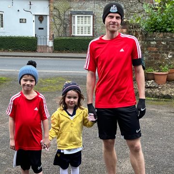 A white man in his 30's, wearing running gear stands on a driveway holding the hand of his toddler daughter, also in running gear. Holding the girl's hand is a young boy, the man's son. All three are smiling and wear woolly hats.