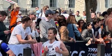 Dave, a white man in his 30s, running the London Marathon, past cheering crowds and trees. He is wearing a white running vest with ClearVision Project on the front