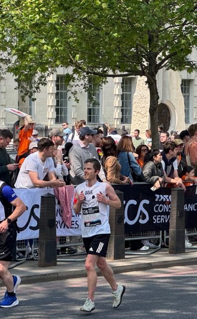 Dave, a white man in his 30s, running the London Marathon, past cheering crowds and trees. He is wearing a white running vest with ClearVision Project on the front