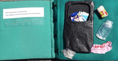 The tactile book Your Help is Needed is open to a page about recycling. A bin contains rubbish, beside it are a plastic bottle, paper wrapper and can ready for recycling. The text in print and braille reads