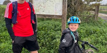 Nate, a seven year old with fair skin and hair stands next to his bike in front of a signboard showing he whole of the Ridgeway Path. Next to him stands his Dad, Dave, a white man in his 30's wearing running kit, stands to his left. Both are smiling.