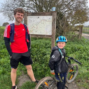 Nate, a seven year old with fair skin and hair stands next to his bike in front of a signboard showing he whole of the Ridgeway Path. Next to him stands his Dad, Dave, a white man in his 30's wearing running kit, stands to his left. Both are smiling.