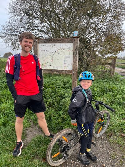Nate, a seven year old with fair skin and hair stands next to his bike in front of a signboard showing he whole of the Ridgeway Path. Next to him stands his Dad, Dave, a white man in his 30's wearing running kit, stands to his left. Both are smiling.