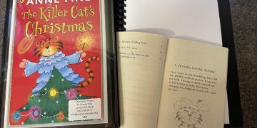 A ClearVision copy of Anne Fine's The Killer Cat's Christmas lies open, showing the text in braille and the print copy of the book open. On the front of the folder is the title and an image of ginger cat in a blue angel costume on top of a Christmas tree.