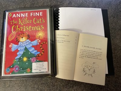 A ClearVision copy of Anne Fine's The Killer Cat's Christmas lies open, showing the text in braille and the print copy of the book open. On the front of the folder is the title and an image of ginger cat in a blue angel costume on top of a Christmas tree.