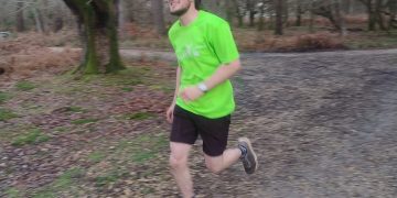 A white man in his 20's with glasses, brown hair and beard runs in a forest. He is smiling and wearing a neon yellow tshirt.