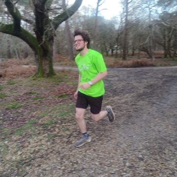 A white man in his 20's with glasses, brown hair and beard runs in a forest. He is smiling and wearing a neon yellow tshirt.