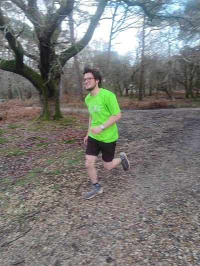 A white man in his 20's with glasses, brown hair and beard runs in a forest. He is smiling and wearing a neon yellow tshirt.