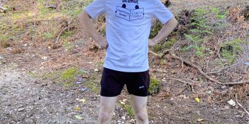 George, a white man in his 20's with brown hair beard and a big smile stands on a woodland path wearing running kit and a white t-shirt with his name and the ClearVision logo.