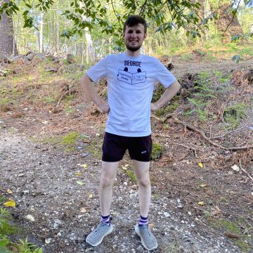 George, a white man in his 20's with brown hair beard and a big smile stands on a woodland path wearing running kit and a white t-shirt with his name and the ClearVision logo.