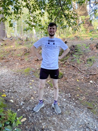 George, a white man in his 20's with brown hair beard and a big smile stands on a woodland path wearing running kit and a white t-shirt with his name and the ClearVision logo.